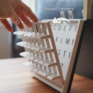 May include: A white and black letter board with a triangular design. The board has rows of white slats and a white surface with black letters. The text "Reset Letters" is visible. A hand is shown interacting with the board.