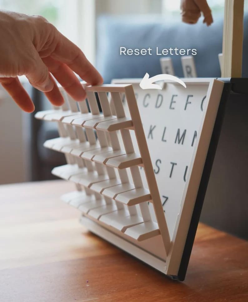 May include: A white and black letter board with a triangular design. The board has rows of white slats and a white surface with black letters. The text "Reset Letters" is visible. A hand is shown interacting with the board.