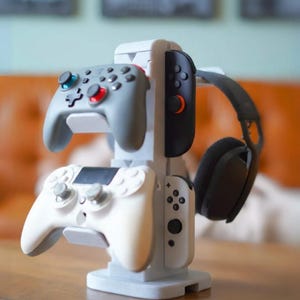 Może przedstawiać: Jasnoszary stojak na kontrolery do gier, na kt&oacute;rym znajdują się dwa szare kontrolery, biały kontroler i czarny zestaw słuchawkowy. Stojak wykonany jest z jasnoszarego tworzywa sztucznego i stoi na drewnianej powierzchni.