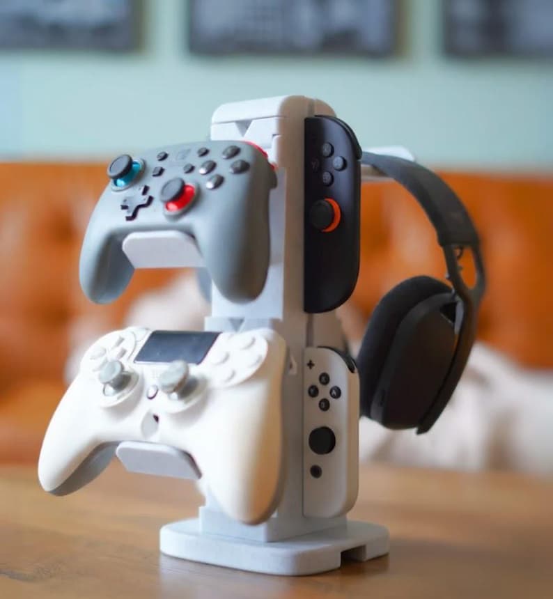 Może przedstawiać: Jasnoszary stojak na kontrolery do gier, na kt&oacute;rym znajdują się dwa szare kontrolery, biały kontroler i czarny zestaw słuchawkowy. Stojak wykonany jest z jasnoszarego tworzywa sztucznego i stoi na drewnianej powierzchni.