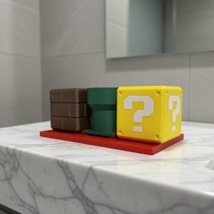 May include: A set of three decorative items: a brown brick block, a green pipe, and a yellow question mark block. The items are arranged on a red base, reminiscent of a video game theme. The question mark block has white question marks on two sides.