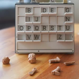 May include: A vintage word game with a white and black frame, featuring letter tiles and small, brown game pieces scattered on a wooden surface. The letters visible are B, D, E, G, I, J, N, O, P, Q, R, S, T, U, V, W, and Z.