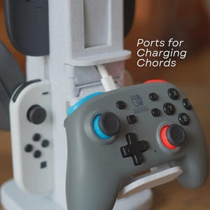 May include: A gray Nintendo Switch controller with blue and red accents rests on a white stand. The stand is part of a larger gray charging station with the text "Ports for Charging Chords". Other controllers are visible.