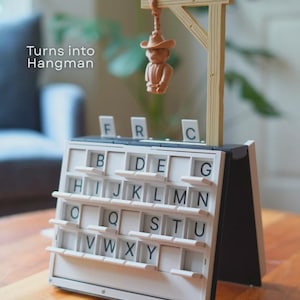 May include: A Hangman game featuring a small figure suspended from a wooden gallows. The game board is white with black letters and a sliding mechanism. The text "Turns into Hangman" is displayed on the left.
