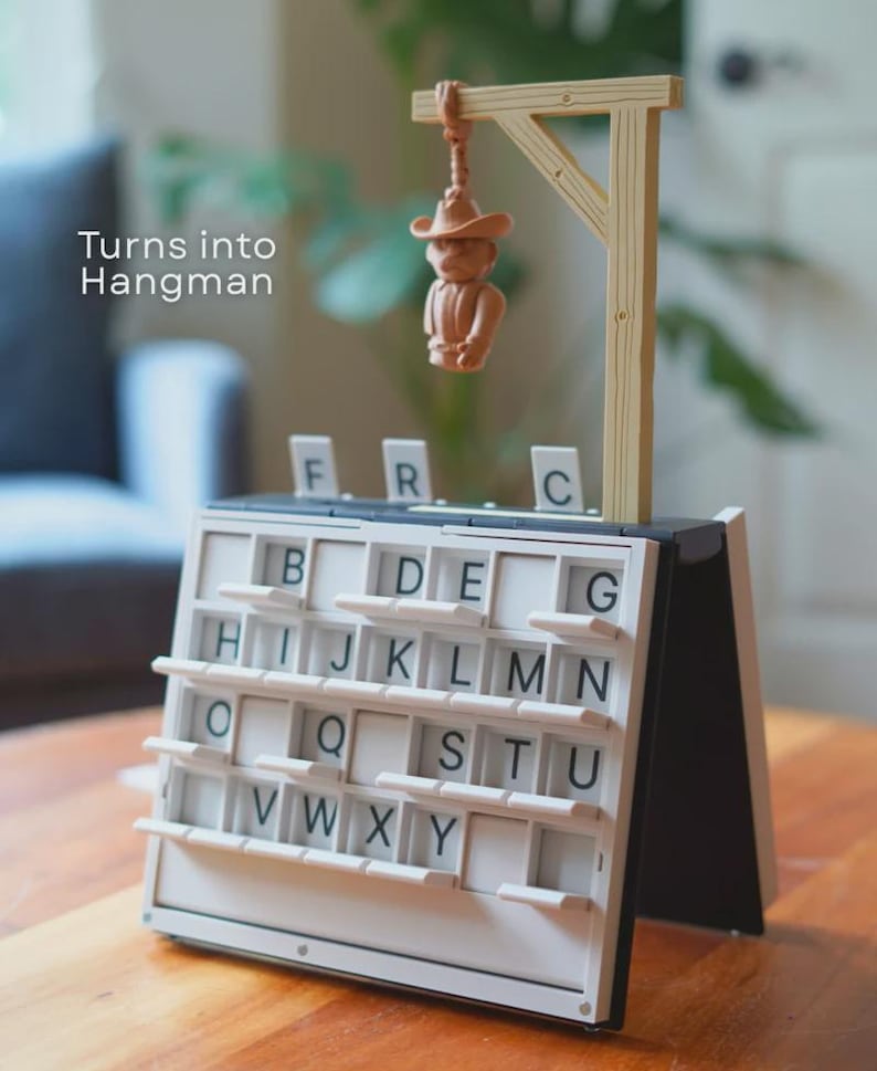 May include: A Hangman game featuring a small figure suspended from a wooden gallows. The game board is white with black letters and a sliding mechanism. The text "Turns into Hangman" is displayed on the left.
