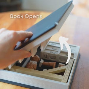 May include: A book with a secret compartment, opened to show miniature items. The book has a dark blue cover and a wooden frame inside. The text "Book Opens" is visible.