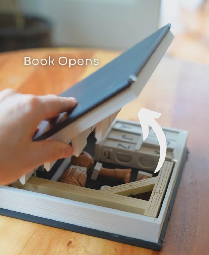 May include: A book with a secret compartment, opened to show miniature items. The book has a dark blue cover and a wooden frame inside. The text "Book Opens" is visible.