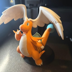 May include: A 3D-printed orange dragon figure with white wings, green details, and a blue sphere on its tail. The dragon is set on a black circular base, with a detailed design. The figurine is a collectible item.