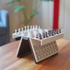 May include: A white and brown portable chess set with a checkered board and chess pieces. The set is on a wooden table. The chess pieces are white and brown. The set has a storage area for the chess pieces.