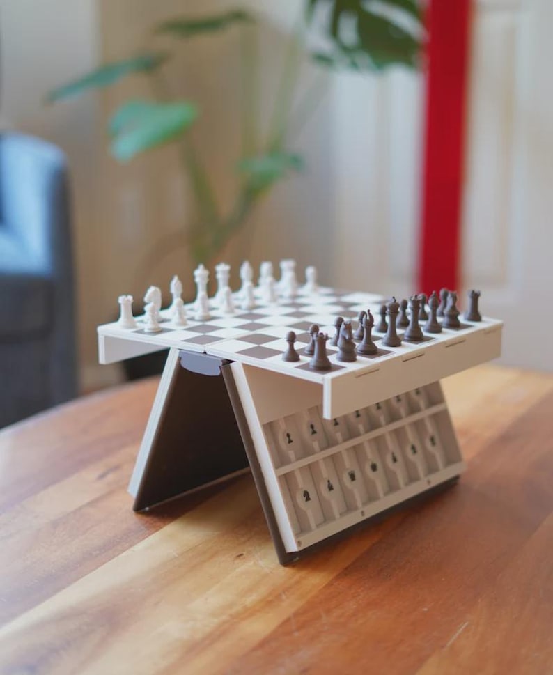 May include: A white and brown portable chess set with a checkered board and chess pieces. The set is on a wooden table. The chess pieces are white and brown. The set has a storage area for the chess pieces.