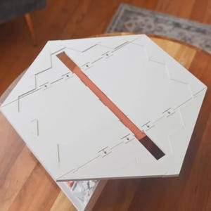 May include: A white, geometric puzzle with a central, rectangular, brown-colored channel. The puzzle is composed of multiple pieces and has a complex, angular design. The object is placed on a wooden surface.