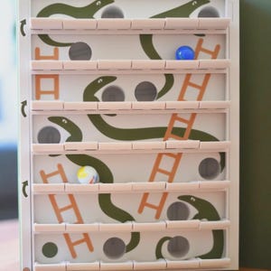 May include: A white and green wooden game with a maze design. The game features green snake shapes, orange ladders, and small holes for marbles. Several colorful marbles are visible within the maze.