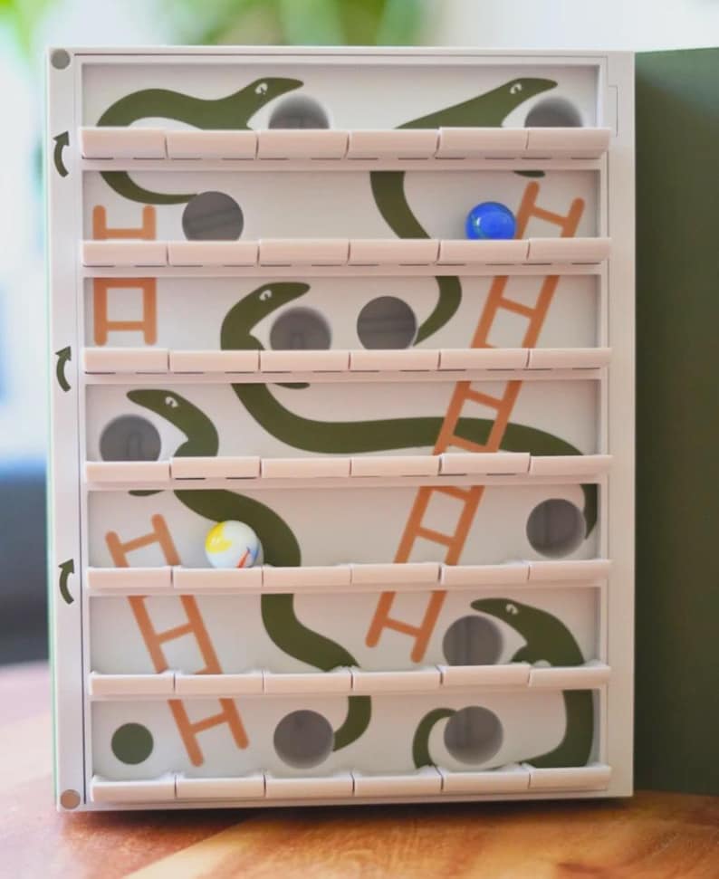 May include: A white and green wooden game with a maze design. The game features green snake shapes, orange ladders, and small holes for marbles. Several colorful marbles are visible within the maze.