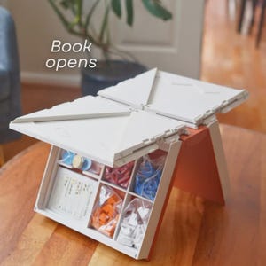 May include: A white and orange board game box, open to reveal game pieces in clear bags and cards. The box is on a wooden table. The text "Book opens" is visible in the image.