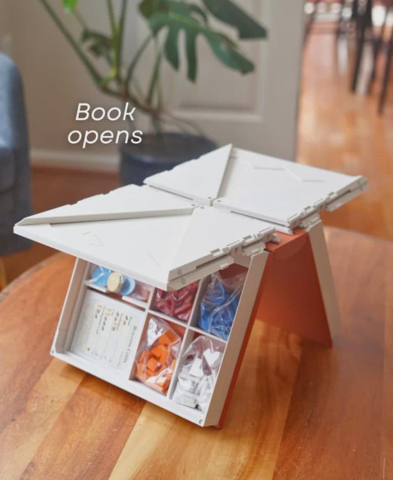 May include: A white and orange board game box, open to reveal game pieces in clear bags and cards. The box is on a wooden table. The text "Book opens" is visible in the image.