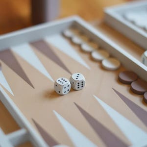 Puede incluir: Un juego de mesa de backgammon con marcas triangulares beige y blancas. Dos dados blancos con puntos negros est&aacute;n sobre el tablero, junto con fichas blancas y marrones. El tablero est&aacute; sobre una superficie de madera.