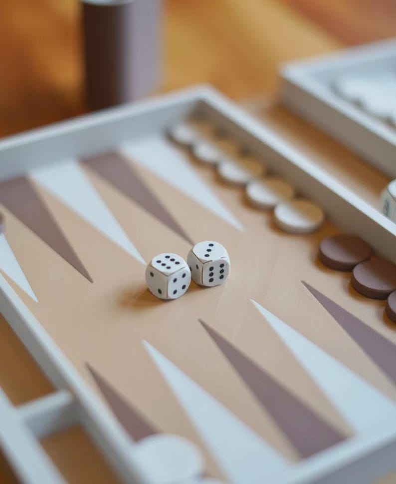 Puede incluir: Un juego de mesa de backgammon con marcas triangulares beige y blancas. Dos dados blancos con puntos negros est&aacute;n sobre el tablero, junto con fichas blancas y marrones. El tablero est&aacute; sobre una superficie de madera.
