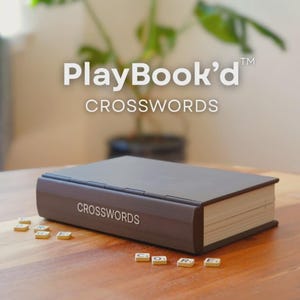 May include: A brown crossword puzzle book with the text "CROSSWORDS" on the side. The book is closed and resting on a wooden surface. Scattered around the book are small letter tiles. The top of the image has the text "PlayBook'd CROSSWORDS".