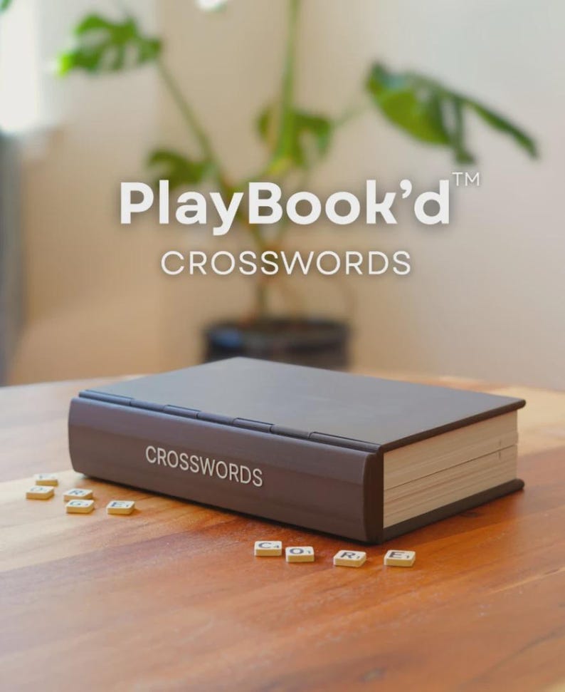 May include: A brown crossword puzzle book with the text "CROSSWORDS" on the side. The book is closed and resting on a wooden surface. Scattered around the book are small letter tiles. The top of the image has the text "PlayBook'd CROSSWORDS".