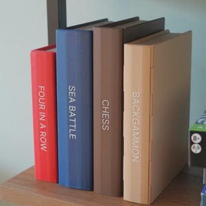 Puede incluir: Cuatro juegos de mesa: "Four in a Row" en rojo, "Sea Battle" en azul, "Chess" en marr&oacute;n y "Backgammon" en beige. Los juegos est&aacute;n colocados verticalmente en una estanter&iacute;a de madera, con los t&iacute;tulos en los lomos.