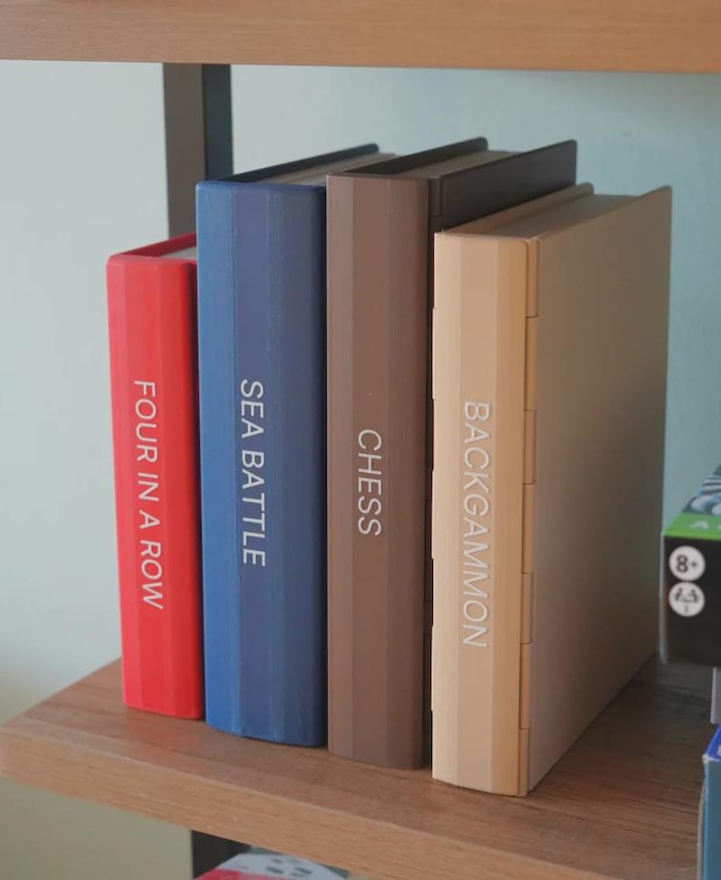 Puede incluir: Cuatro juegos de mesa: "Four in a Row" en rojo, "Sea Battle" en azul, "Chess" en marr&oacute;n y "Backgammon" en beige. Los juegos est&aacute;n colocados verticalmente en una estanter&iacute;a de madera, con los t&iacute;tulos en los lomos.