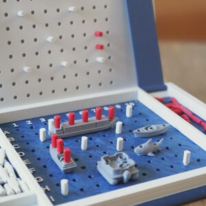 Op de afbeelding: Battleship bordspel met een blauw speelveld, grijze schepen en rode en witte pinnen. Het spel zit in een witte en blauwe koffer, met pinnen opgeborgen aan de zijkant.