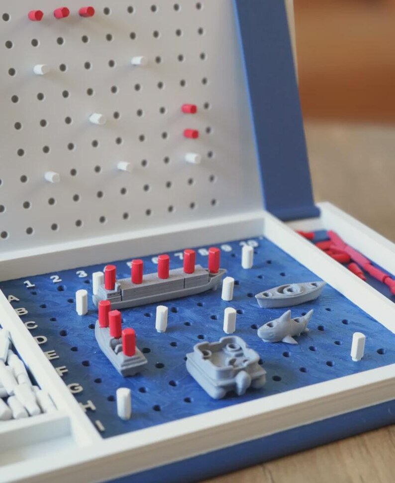 Op de afbeelding: Battleship bordspel met een blauw speelveld, grijze schepen en rode en witte pinnen. Het spel zit in een witte en blauwe koffer, met pinnen opgeborgen aan de zijkant.