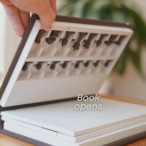 May include: A brown and white book-shaped box with chess pieces stored inside. The box is open, revealing the chess pieces and the text "Book opens". The box is on a wooden surface.