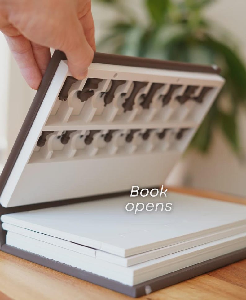 May include: A brown and white book-shaped box with chess pieces stored inside. The box is open, revealing the chess pieces and the text "Book opens". The box is on a wooden surface.