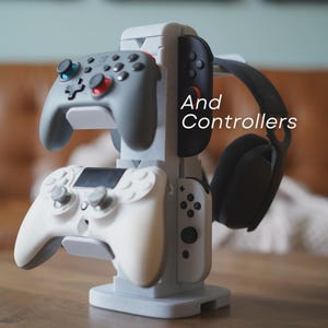 Może przedstawiać: Szary stojak na kontrolery do gier, na kt&oacute;rym znajdują się dwa kontrolery, para słuchawek i mały kontroler. Na stojaku widnieje napis "And Controllers". Kontrolery są szare i białe.