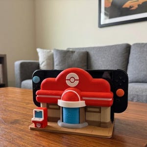 May include: A red and white decorative stand designed to hold a black Nintendo Switch. The stand is shaped like a building with a red roof and a white logo. A small red arcade machine is on the left.
