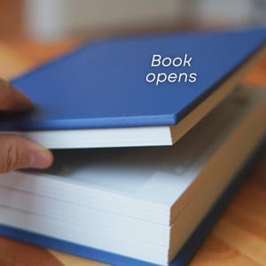 Op de afbeelding: Een blauw hardcover boek met de tekst "Book opens" wordt gedeeltelijk geopend getoond. De pagina's van het boek zijn wit en de kaft is effen blauw. Het boek ligt op een houten oppervlak.