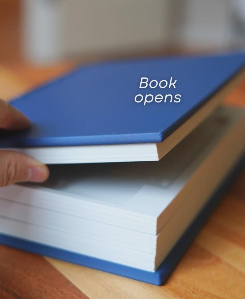 Op de afbeelding: Een blauw hardcover boek met de tekst "Book opens" wordt gedeeltelijk geopend getoond. De pagina's van het boek zijn wit en de kaft is effen blauw. Het boek ligt op een houten oppervlak.