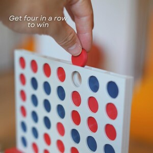 Może przedstawiać: Zbliżenie gry Connect Four. Plansza do gry jest biała z czerwonymi i niebieskimi okrągłymi elementami. Ręka umieszcza czerwony element w planszy. Tekst na obrazku brzmi: "Ustaw cztery w rzędzie, aby wygrać."