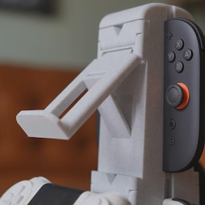 Może przedstawiać: Jasnoszary stojak na konsolę do gier z wbudowanym ramieniem podtrzymującym. Stojak jest przeznaczony do trzymania kontrolera konsoli do gier, z dołączonym czarno-szarym kontrolerem. Stojak ma teksturowaną powierzchnię.