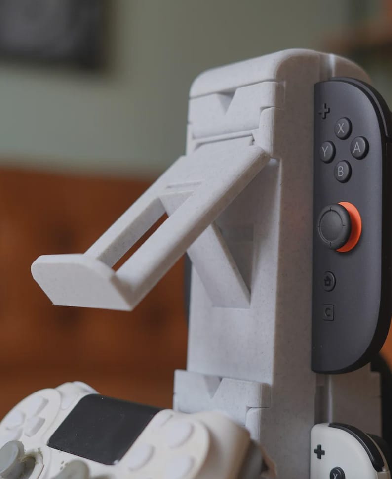 Może przedstawiać: Jasnoszary stojak na konsolę do gier z wbudowanym ramieniem podtrzymującym. Stojak jest przeznaczony do trzymania kontrolera konsoli do gier, z dołączonym czarno-szarym kontrolerem. Stojak ma teksturowaną powierzchnię.