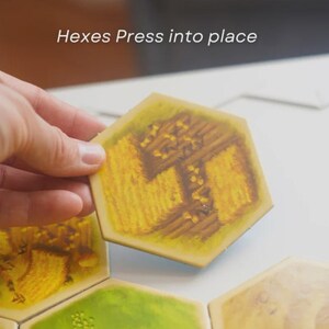 May include: A hand holding a yellow hexagonal tile with a wheat field design. The tile is part of a board game, with the text "Hexes Press into place" visible. Other tiles with different designs are also present.
