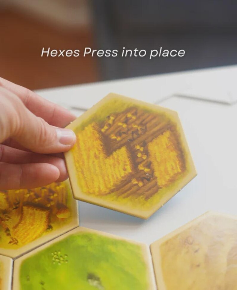 May include: A hand holding a yellow hexagonal tile with a wheat field design. The tile is part of a board game, with the text "Hexes Press into place" visible. Other tiles with different designs are also present.