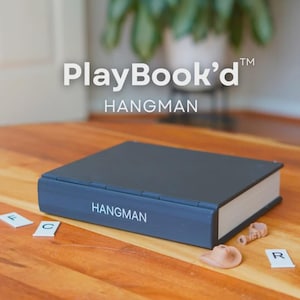 May include: A dark blue PlayBook'd Hangman game set on a wooden surface. The game is designed to look like a book. The word "HANGMAN" is printed in white on the side of the book. Scattered around the book are game pieces.