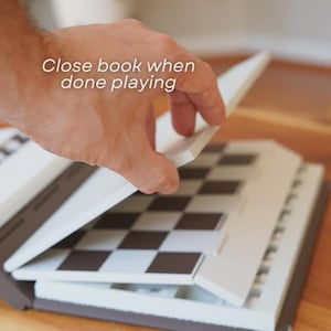 May include: A portable chess set with a brown and white exterior. The chess board is black and white. The text "Close book when done playing" is visible on the top of the image.