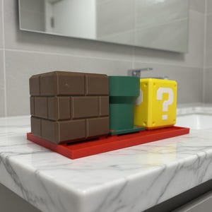 May include: A red tray holds a brown brick block, a green pipe, and a yellow question mark block. The items are arranged on a white marble countertop in a bathroom setting. The objects are reminiscent of a popular video game.