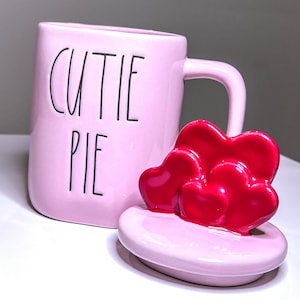 May include: A pink ceramic mug with the words "CUTIE PIE" printed on the side. The mug has a matching pink lid topped with three red heart-shaped decorations. The mug has a curved handle.