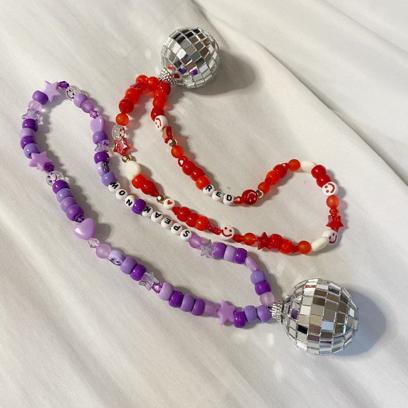 May include: Two beaded necklaces with purple and red beads, silver disco ball charms, and letter beads that spell "Speak Now" and "Red".