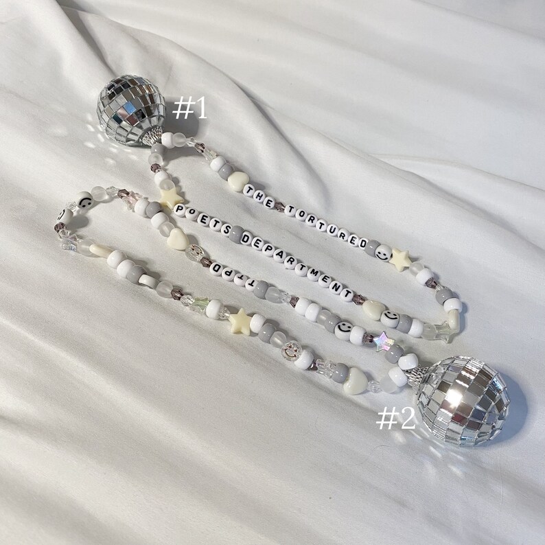 May include: Two silver disco ball phone charms with a beaded chain. The chain has white and gray beads with the words "The Tortured Poets Department" on it.