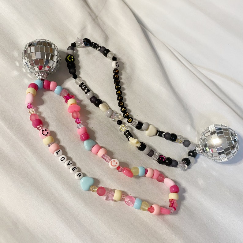 May include: Two beaded necklaces with disco ball charms. One necklace is black with white beads and the word "REPUTATION" spelled out. The other necklace is pink and white with the word "LOVER" spelled out.