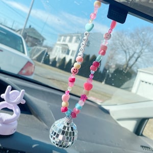 May include: A pink, blue, and yellow beaded car charm with a silver disco ball hanging from it. The beads spell out the word "LOVER" and include smiley faces, hearts, and stars.