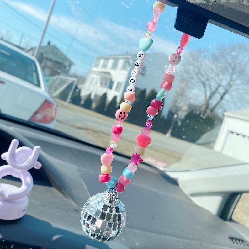 May include: A pink, blue, and yellow beaded car charm with a silver disco ball hanging from it. The beads spell out the word "LOVER" and include smiley faces, hearts, and stars.