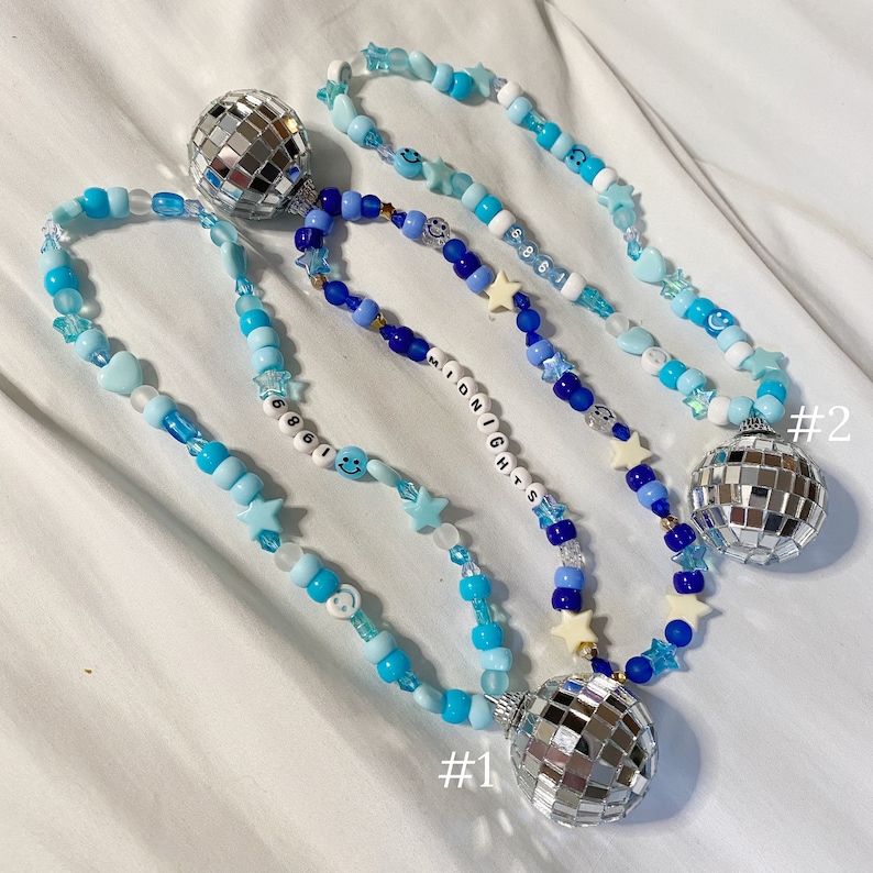 May include: Three beaded necklaces with disco ball charms. The necklaces are made with blue and white beads, including stars, smiley faces, and the word "MIDNIGHTS". The necklaces are labeled #1 and #2.