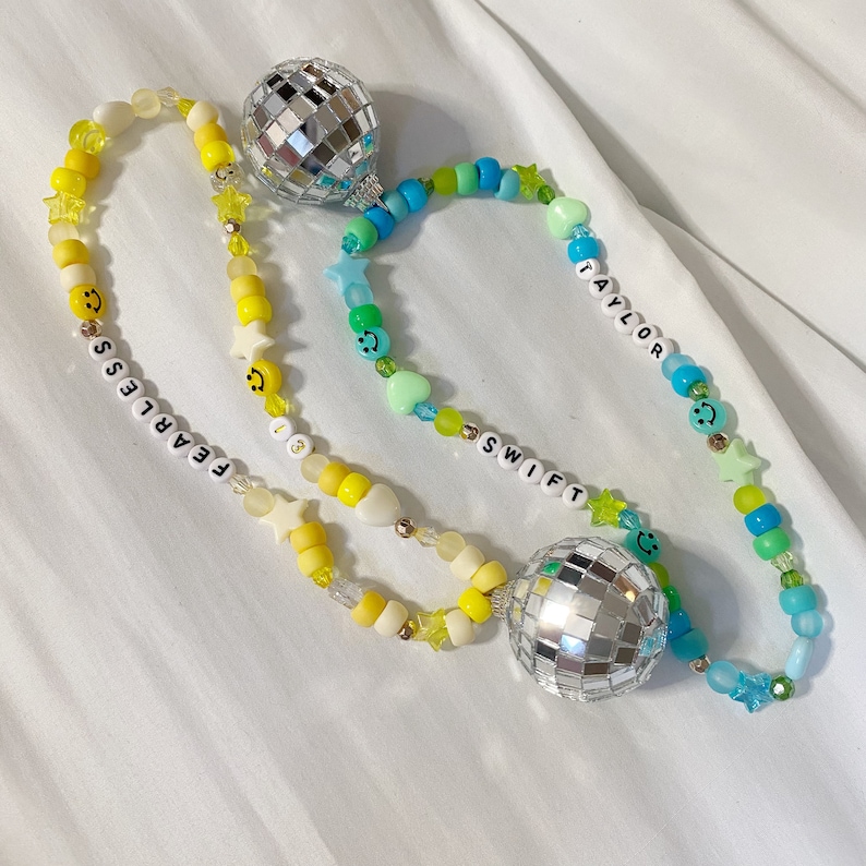 May include: A beaded necklace with two disco ball charms. The beads are yellow, blue, green, and white. The necklace has the words "Fearless" and "13" on one side and "Taylor Swift" on the other.