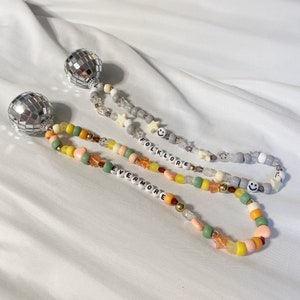 May include: Two beaded necklaces with silver disco ball charms. The necklaces are made with grey, green, yellow, and orange beads. One necklace has the word "folklore" spelled out in white beads and the other has the word "evermore" spelled out in white beads.